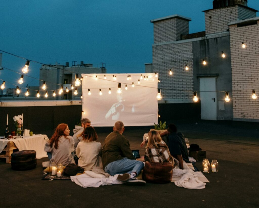 A group of friends enjoys a rooftop movie night under string lights with a projector and cozy blankets.